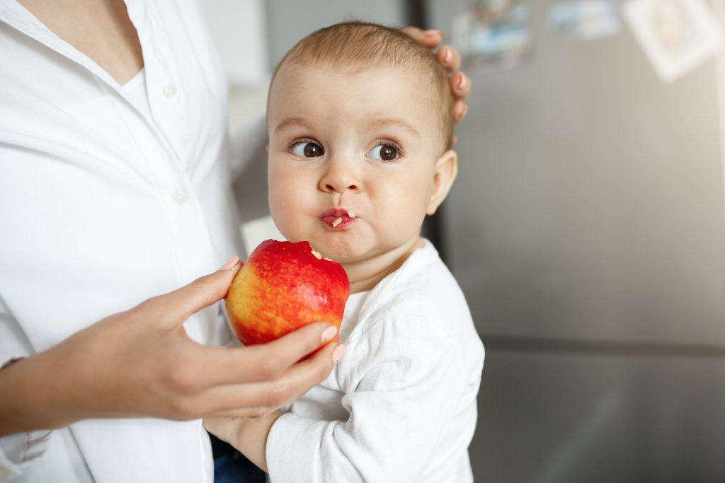 Close up of cute little baby with big brown eyes and funny expression eating apple from mother hands in light cozy kitchen.