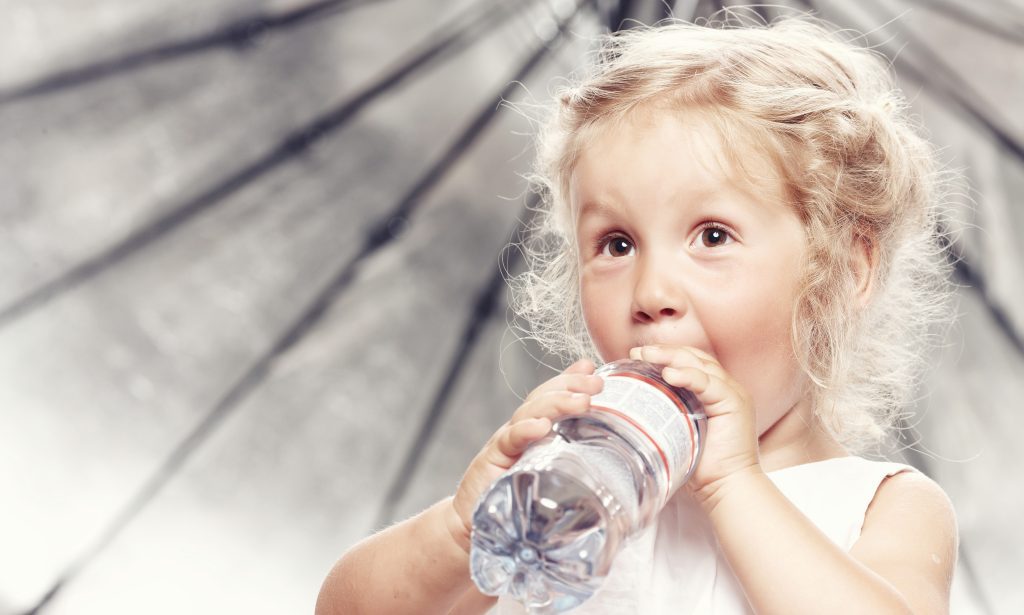 Portrait of a funny cute little girl in a casual dress, drink water while sitting in studio.