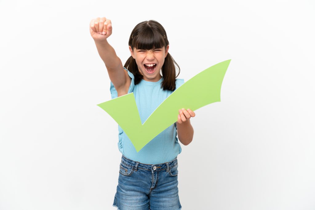 Little caucasian kid isolated on white background holding a check icon and celebrating a victory
