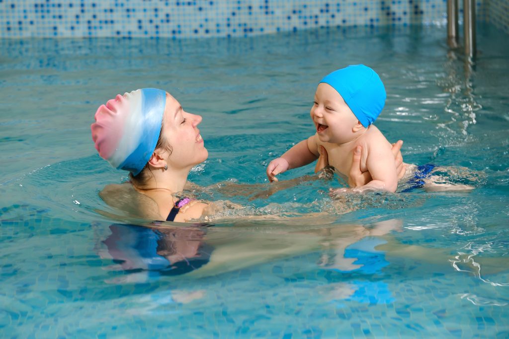 Baby with mother trainer in indoor swimming pool having lesson